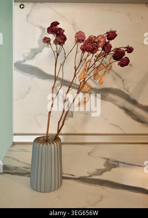 Eine vertikale Nahaufnahme mit Fokus auf einer kleinen Keramikvase mit trockenen, detaillierten Pflanzenstoffen auf einer marmorstrukturierten Oberfläche. Stockfoto
