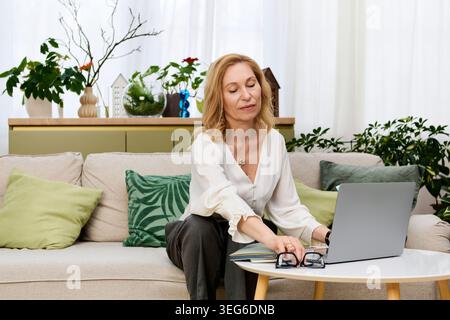 Eine schöne, reife Frau sitzt friedlich und konzentriert sich auf ihren Laptop in einem stilvollen, gemütlichen Wohnzimmer. Stockfoto
