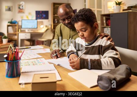 Schwarzer Vater hilft dem Sohn im Schulalter, Hausaufgaben am Tisch zu schreiben, unterstützt das Lernen zu Hause und die Einbeziehung der Eltern. Ideal für Bildung, Nachhilfe, Erziehung, Kinderentwicklungskampagnen Stockfoto