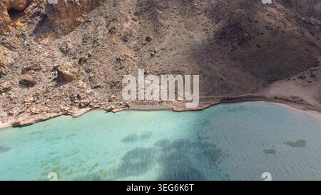 Aus der Vogelperspektive auf eine felsige Wüstenküste trifft auf klares türkisfarbenes Wasser in einer abgeschiedenen Bucht und zeigt karge Hügel und flache Küsten-Lagunen-Strukturen. Also Stockfoto