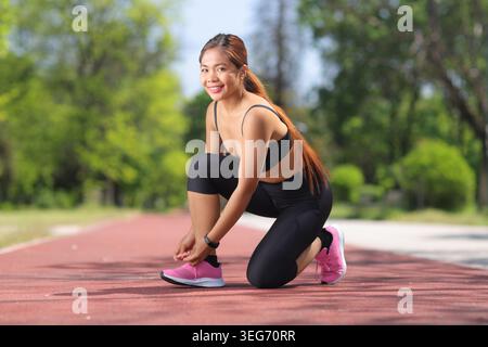 Junge Fitnessfrau kniet draußen auf einer Laufbahn und bindet ihre Schnürsenkel Stockfoto