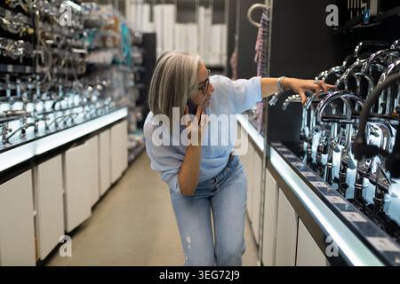 Eine Frau mit grauem Haar inspiziert Küchenarmaturen in einem gut beleuchteten Haus-Baumarkt. Sie hält ihr Telefon am Ohr, während sie Optionen vergleicht und moderne Armaturen präsentiert. Stockfoto