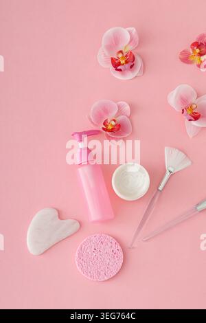 Trendige Selbstpflege- und Hautpflegekomposition mit Gesichtscreme, Kosmetikflasche, Schönheitszubehör und Orchideenblüten auf pastellrosa Hintergrund. Stockfoto