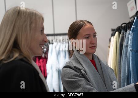 Zwei Frauen stöbern in einer hellen Boutique, vergleichen Kleidungsstücke auf einem Regal und halten Kleidung hoch, während sie gemeinsam in einem modernen Einzelhandelsgeschäft einkaufen und Casua genießen Stockfoto