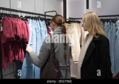 Zwei Frauen stöbern in einer hellen Boutique, vergleichen Kleidungsstücke auf einem Regal und halten Kleidung hoch, während sie gemeinsam in einem modernen Einzelhandelsgeschäft einkaufen und Casua genießen Stockfoto