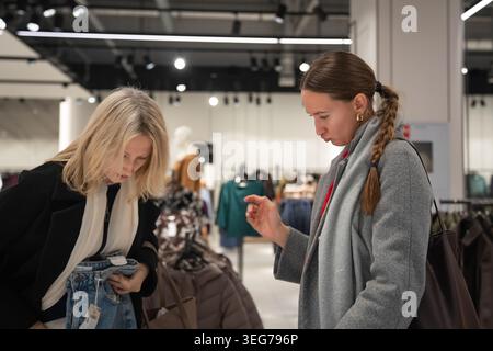 Zwei Frauen stöbern in einer hellen Boutique, vergleichen Kleidungsstücke auf einem Regal und halten Kleidung hoch, während sie gemeinsam in einem modernen Einzelhandelsgeschäft einkaufen und Casua genießen Stockfoto