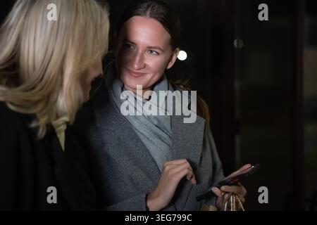 Zwei Frauen, die nachts draußen stehen, sich unterhalten und lächeln, während sie Inhalte auf einem Smartphone ansehen und Einkaufstaschen in der Hand halten Stockfoto