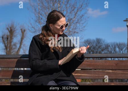 Frau, die an einem sonnigen Tag im Freien ein Buch liest, sich auf den Text konzentriert, Sonnenbrille mit Katzenaugen und einen schwarzen Mantel trägt, einen Moment des Studiums oder der Freizeit genießt Stockfoto