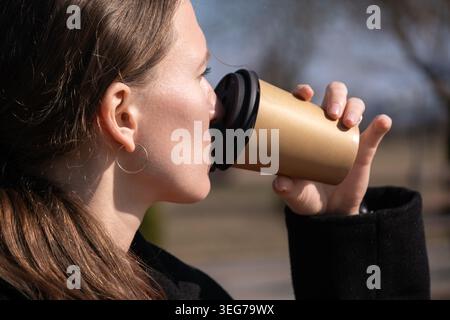 Eine junge Frau mit braunem Haar schlürft ein warmes Getränk aus einer nachhaltigen, wiederverwendbaren Kaffeetasse zum Mitnehmen und genießt eine erfrischende Morgenpause draußen auf einem Stockfoto