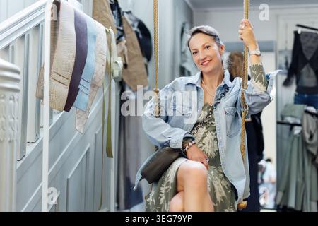 Porträt junger Erwachsener hübsch glücklich lächelnde Frau genießt es, drinnen auf Vintage Schaukel zu sitzen. Weibliche Person in lässigem Kleid Glücksausdruck Stockfoto