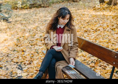 Die Frau sitzt auf einer Holzbank im Park und legt Weissagungskarten auf den Sitz. Tarotkartenlesung, moderne spirituelle Praktiken, esoterisches Lebensstil-Ritual Stockfoto
