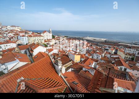 Panoramablick auf Alfamas rot gekachelte Dächer, die in Richtung des Tejo kaskadieren und Lissabons historische Architektur, enge Gassen und die Küste zeigen Stockfoto