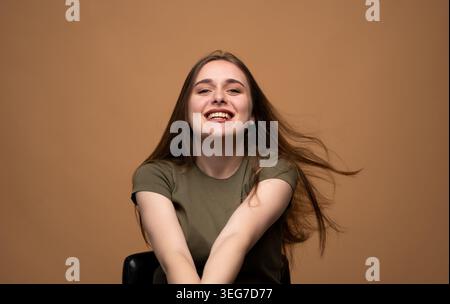 Junge Frau mit einem olivgrünen T-Shirt, die glücklich in die Kamera blickt, ihr langes Haar fließend, zeigt echte Freude und positive Emotionen Stockfoto