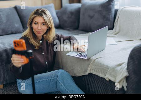 Frau, die am Online-Kurs und an einem virtuellen Meeting auf dem Smartphone teilnimmt, während sie von zu Hause aus mit einem Laptop studiert. Hochwertige Fotos Stockfoto