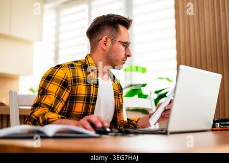 Ein junger Mann in Brille sitzt an einem Holztisch, konzentriert sich auf Dokumente, während er für freiberufliche Arbeit mit dem Laptop arbeitet. Stockfoto
