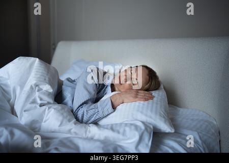 Zufriedene, ruhige Frau mittleren Alters, die sich in einem gemütlichen Bett entspannt und sich zu Hause entspannt und den langsamen Morgen genießt. Stockfoto