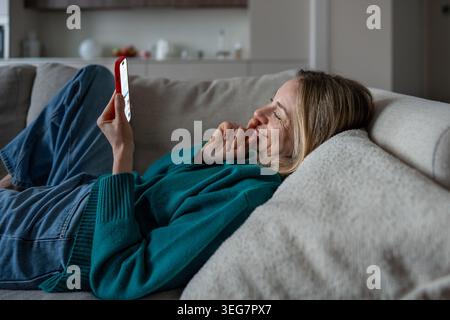 Lächelnde Frau mittleren Alters, die sich mit dem Telefon auf der Couch entspannt, im Internet surft, Online-Nachrichten liest Stockfoto