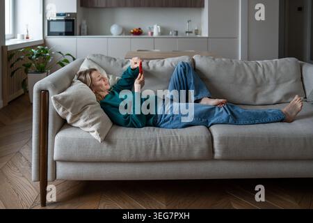 Fröhliche Frau mittleren Alters, die auf der Couch in einer gemütlichen Wohnung ruht, eingetaucht in aufmunternde Inhalte am Telefon. Stockfoto