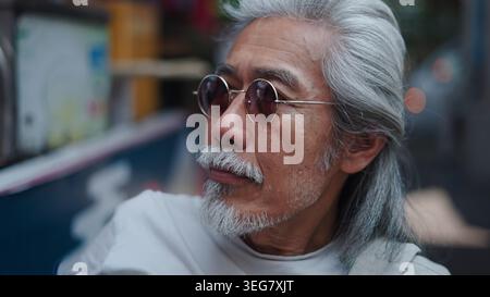 Ein älterer asiatischer Mann mit langen grauen Haaren und Bart, der eine Sonnenbrille trägt und nachdenklich aussieht Stockfoto