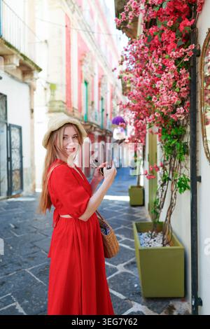 Modische Reisende erkunden die farbenfrohe europäische Altstadt mit Smartphone und floralem Hintergrund an sonnigen Tagen. Stockfoto