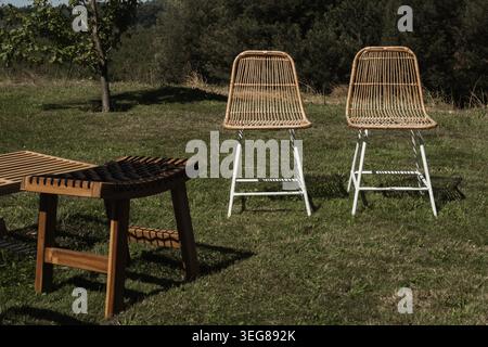 Rattanstühle stehen auf einem grasbewachsenen Feld. Holzhocker in der Nähe sorgen für rustikalen Charme. Sonnenlicht wirft Schatten auf grünen Rasen. Die Natur ist ruhig und im Freien Stockfoto
