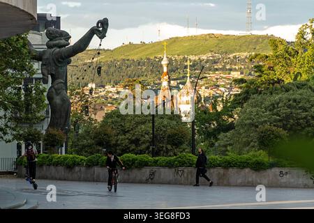 Eine Statue steht im Vordergrund, während die Menschen einen gepflasterten Weg in der Nähe eines grünen Hügels in Tiflis, Georgien, entlang gehen. Stockfoto