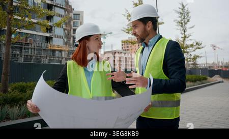 Kaukasier zwei Kollegen Baumeister Architekten Frau Mann diskutieren Blueprint Plan Kollegen Ingenieure Frau männlich in Helmen Hardhelme Westen Papier Dokument Stockfoto