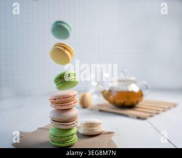 Luftige, pastellfarbene Szene mit gestapelten und schwebendem Macarons in weichem natürlichem Licht auf einer weißen Tischplatte, mit einer verschwommenen Glas-Teekanne mit Bernsteintee A Stockfoto