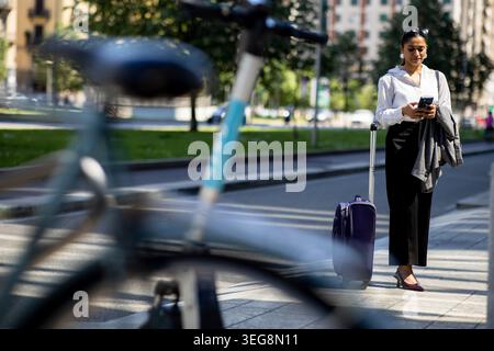 Eine Frau steht auf einer Stadtstraße mit einem Koffer neben sich. Sie schaut auf ihr Handy, während ein Fahrrad in der Nähe parkt. Es ist tagsüber und es gibt bui Stockfoto
