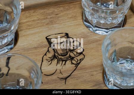 Nahaufnahme von transparenten Shot-Gläsern auf einem hölzernen Serviertablett mit einer gravierten Hand, die eine Glasillustration hält. Stockfoto