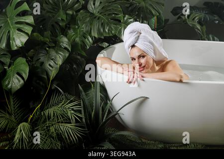 Eine ruhige Frau genießt einen entspannenden Moment in einer weißen Badewanne, umgeben von tropischen Pflanzen, die Ruhe und Selbstpflege in einer Spa-ähnlichen Umgebung bietet. Stockfoto