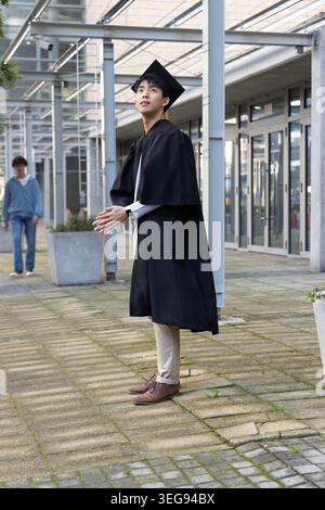 Asiatischer Mann, der unter Pergola im Campus-Hof steht, in schwarzem Abschlusskleid und Mortarboard Stockfoto