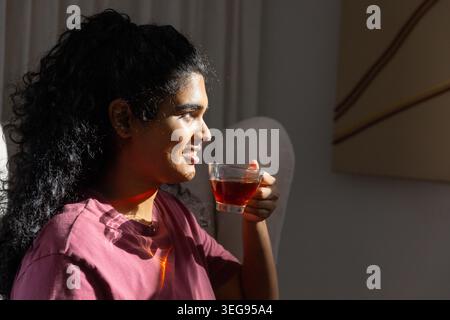 Indische Frau, die im Wohnzimmer sitzt und eine Tasse Tee aus klarem Glas hält, kastanienbraunes T-Shirt, Kopierraum Stockfoto