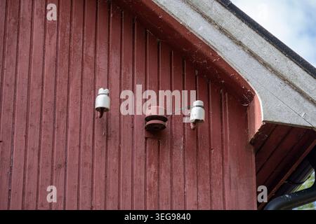 Alte alte elektrische Keramikisolatoren mit fehlenden Drähten, die unter der Dachlinie an einem roten Holzhaus befestigt sind Stockfoto