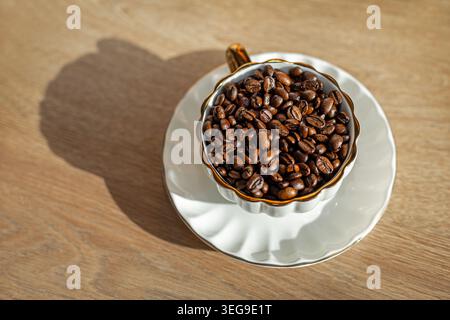 Geröstete Kaffeebohnen in eleganter Tasse. Geröstete Kaffeebohnen füllen eine weiße Tasse auf einer Untertasse, die von warmem Sonnenlicht auf einem Holztisch beleuchtet wird. Stockfoto