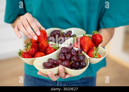 Frische Erdbeeren und Trauben auf Servierplatte. Person, die ein geteiltes Obsttablett mit frischen Erdbeeren und roten Trauben hält, perfekt für gesunde Stockfoto