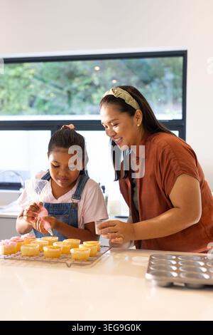 Asiatische Frau, die Mädchen in Denim-Overalls führt, die Cupcakes auf der Küchentheke mit Paspelbeutel dekoriert Stockfoto