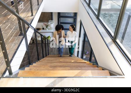 Zwei Erwachsene Frauen, die im Loft eine Holztreppe hinaufgehen, Blazer tragen und gerollte Dokumente halten Stockfoto