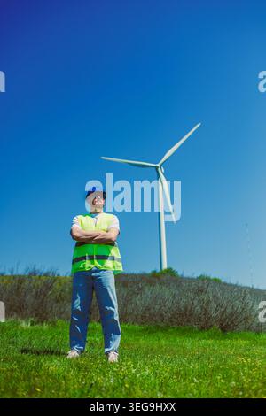 Ein Windmühle-Ingenieur in einer Sicherheitsweste steht mit überkreuzten Armen auf einem grasbewachsenen Feld. Hinter ihm steht eine große Windturbine vor blauem Himmel. Es ist tagsüber A Stockfoto
