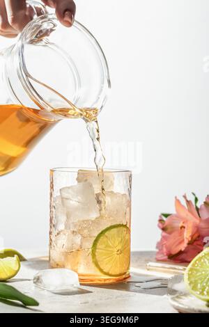 Kalter Tee in ein Glas mit Limettenscheibe und Eiswürfeln auf weißem Hintergrund gießen. Zubereitung von erfrischendem Eisgetränk Stockfoto