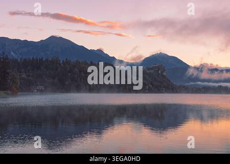 043 rötlicher, nebeliger Sonnenaufgang über dem Bleder See und der Burg mit den Karawanken (Karavanke) im Hintergrund, vom Velika Zaka Pier aus gesehen. Bled-Slowenien Stockfoto