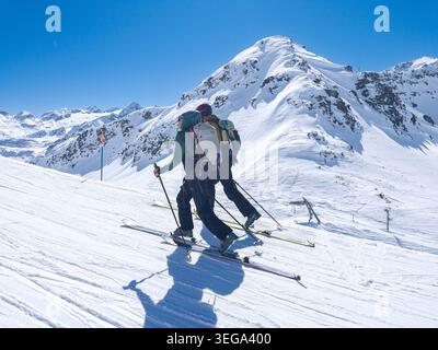 Zwei Skibergsteiger, die an einem hellen, sonnigen Tag in La Thuile auf einer unberührten, schneebedeckten Piste vor der Kulisse majestätischer Berggipfel bergauf fahren, Stockfoto
