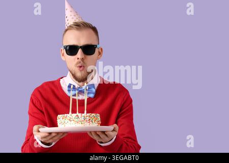 Junger Mann, der Kerzen auf Geburtstagskuchen vor violettem Hintergrund ausbläst Stockfoto