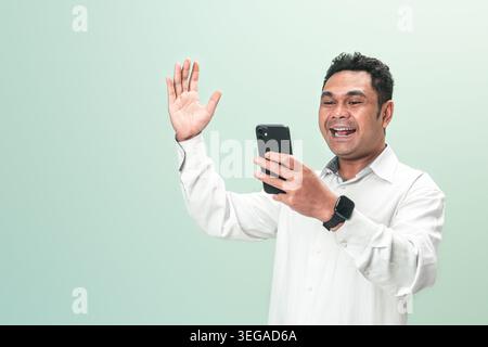 Ein glücklicher Indonesier in einem weißen Hemd lächelt, während er während eines Videoanrufs auf seinem Mobiltelefon mit der Hand winkt vor einem grünen Hintergrund. Stockfoto