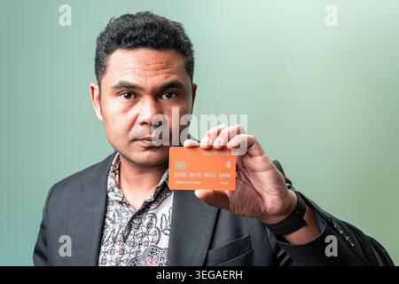 Ein ernster indonesischer Geschäftsmann in einem Blazer zeigt ein orangefarbenes Kreditkarten-Mockup, passend für Finanz-, Digitalzahlungs- und Diversitätskonzepte. Stockfoto