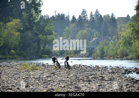 Malerische Sommerlandschaft mit zwei Mountainbikes, die an einem Kiesstrand am Elbow River in Calgary, Alberta, geparkt werden. Im Hintergrund sind die Menschen en Stockfoto