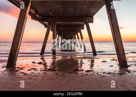 Sonnenuntergang unter dem Glenelg Jetty mit Reflexionen auf nassem Sand in Südaustralien Stockfoto