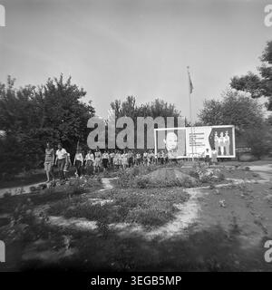 Sloviansk, 1983, Slavkurort, Pionierlager Wishnegorsk. Eine Gruppe von Kindern marschiert an einem Sommertag auf einem gepflasterten Weg durch den grünen Campingplatz. Hinter ihnen steht eine große ideologische Werbetafel mit einem Porträt von Wladimir Lenin und einem kommunistischen Jugendslogan neben einem hohen Fahnenmast. Ein historisches Schwarzweiß-Archivfoto, das die authentische Atmosphäre der Epoche, die organisierten Freizeitaktivitäten, die ideologische Umgebung und den friedlichen Donbass feststellt. Stockfoto
