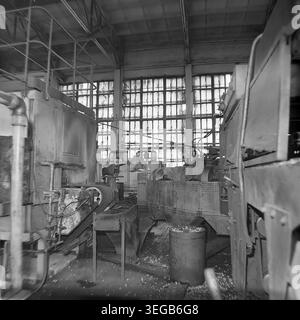 Sloviansk, 1981, AIZ (Armature-Insulator Plant). Eine leere Ansicht der Fabrikhalle mit schweren Industriemaschinen, einem Fördermechanismus und einem Metallbehälter, der mit entsorgtem Material gefüllt ist. Sonnenlicht strömt durch eine massive Wand aus mehrverglasten Industriefenstern im Hintergrund und beleuchtet den Arbeitsbereich. Ein historisches Schwarzweiß-Archivfoto, das die authentische Atmosphäre der Epoche, die Größe der Produktionsanlagen und die industrielle Umgebung feststellt. Ein detaillierter Einblick in die Vergangenheit, die sowjetische Industrie und den friedlichen Donbass. Stockfoto