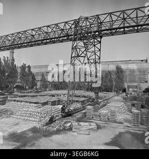 Sloviansk, 1981, AIZ (Armature-Insulator Plant). Eine breite Außenansicht mit einem massiven Portalkran aus Metall, der über einem Fabrikhof thront. Der Hof ist komplett gefüllt mit ordentlich angeordneten Stapeln fertiger Keramikisolatoren und hölzernen Transportkisten, mit hohen Bäumen im Hintergrund. Ein historisches Schwarzweiß-Archivfoto, das die authentische Atmosphäre der Epoche, die enorme Größe der industriellen Logistik und das Außendesign der Fabrik feststellt. Ein detaillierter Einblick in die Vergangenheit, die sowjetische Industrie und den friedlichen Donbass. Stockfoto